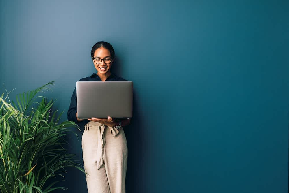 A woman wearing glasses and a dark shirt stands by a green plant, smiling while holding and looking at an open laptop against a plain teal wall.