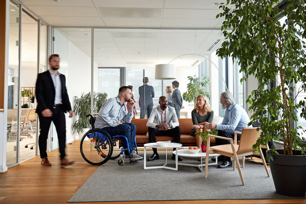 A diverse group of five people, including a man in a wheelchair, are having a meeting in a modern office lounge. Another person is walking by. The area is bright with large windows, plants, and contemporary furniture.