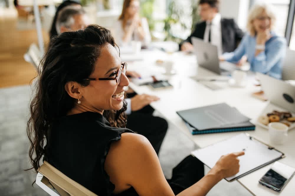 A woman smiling and holding a pen sits at a conference table with colleagues during a meeting. Laptops, papers, and coffee cups are on the table, and people are engaged in conversation in a bright office.
