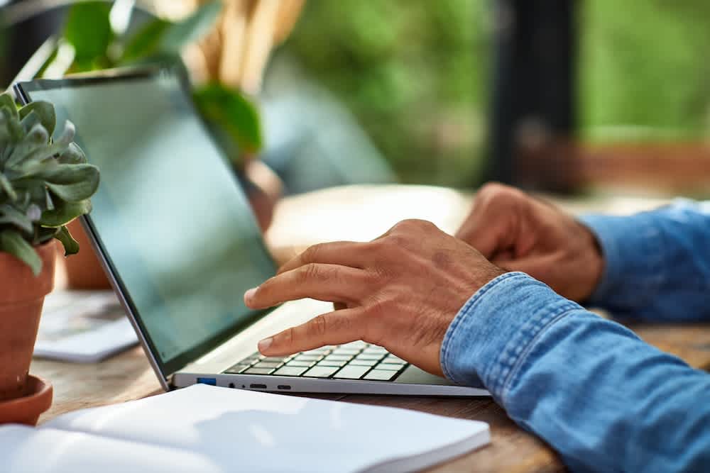 A person in a denim shirt types on a laptop at a wooden table, surrounded by potted plants and an open notebook, in a bright, natural setting.