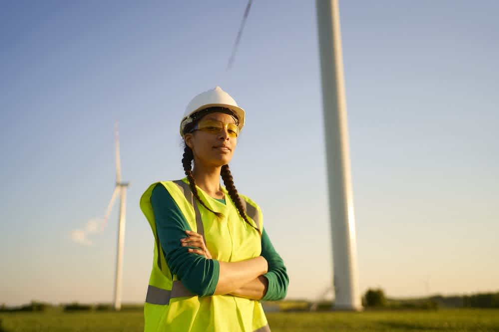 A person wearing a safety helmet, reflective vest, and protective glasses stands with arms crossed in front of wind turbines on a sunny day, representing work in renewable energy.