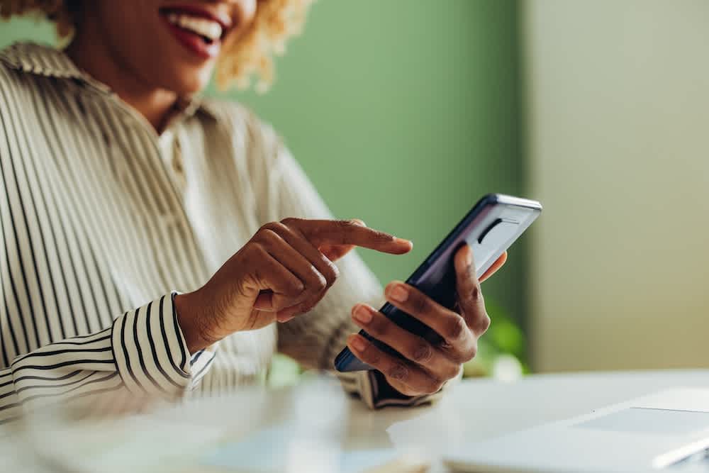 A smiling person wearing a striped shirt sits at a desk and uses a smartphone, holding it in one hand and tapping the screen with the other. The background is softly focused and green.