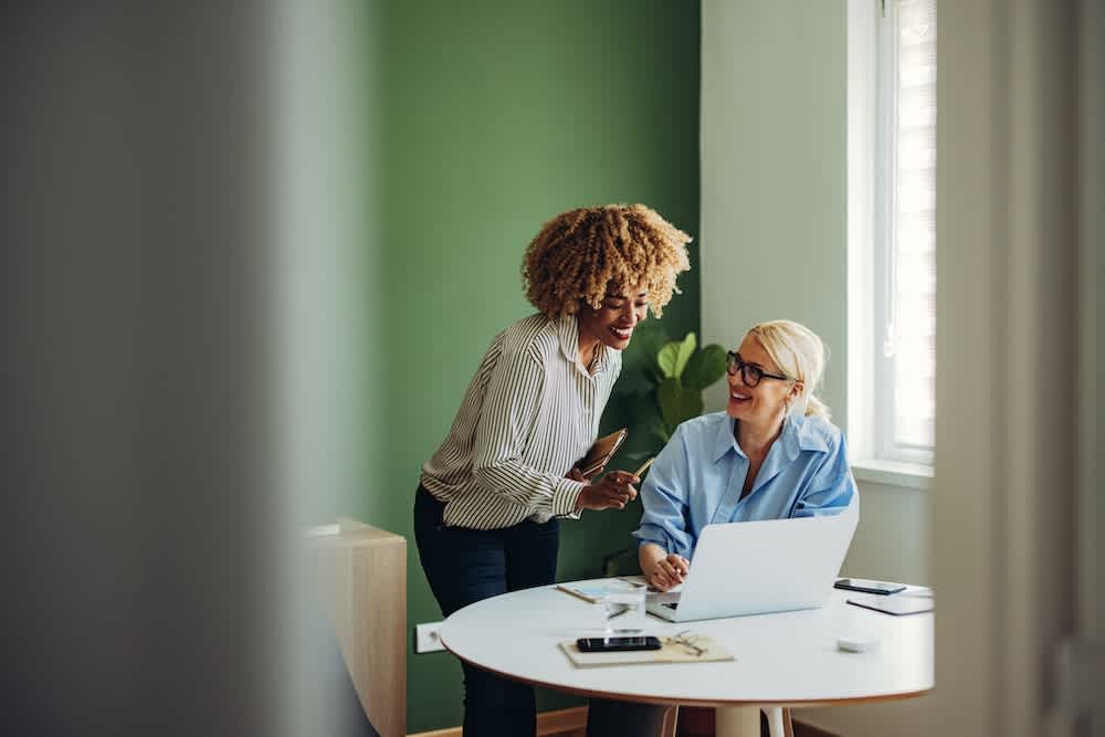 Two women in an office smile and discuss work at a round desk with a laptop, notebook, and smartphone. One woman is sitting and the other is standing, holding a tablet. Sunlight enters through a nearby window.