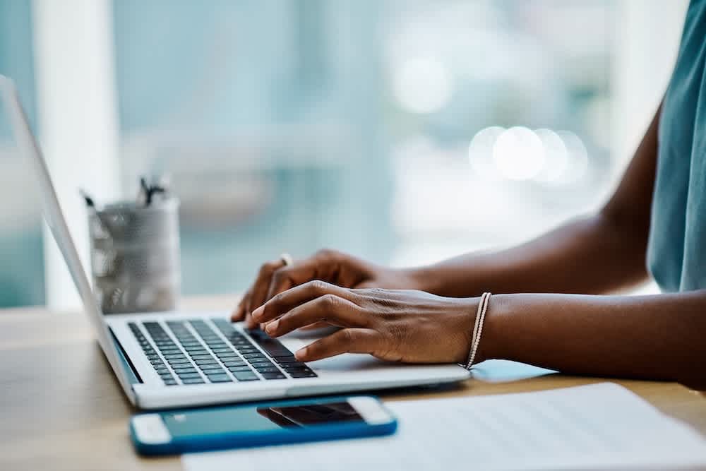 A person typing on a laptop at a desk, with a smartphone and papers nearby. The person is wearing a bracelet and a ring, and the setting appears to be a bright, modern workspace.