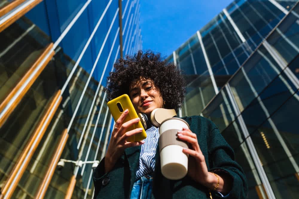 A person with curly hair stands outside modern glass buildings, smiling while looking at a yellow smartphone. They wear headphones around their neck and hold a reusable coffee cup.