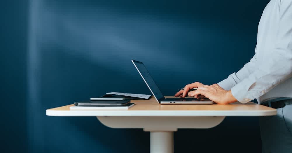 A person in a light shirt stands at a modern desk, typing on a laptop. Notebooks and a tablet rest nearby, with a dark blue background behind them. The image conveys a professional, focused work environment.