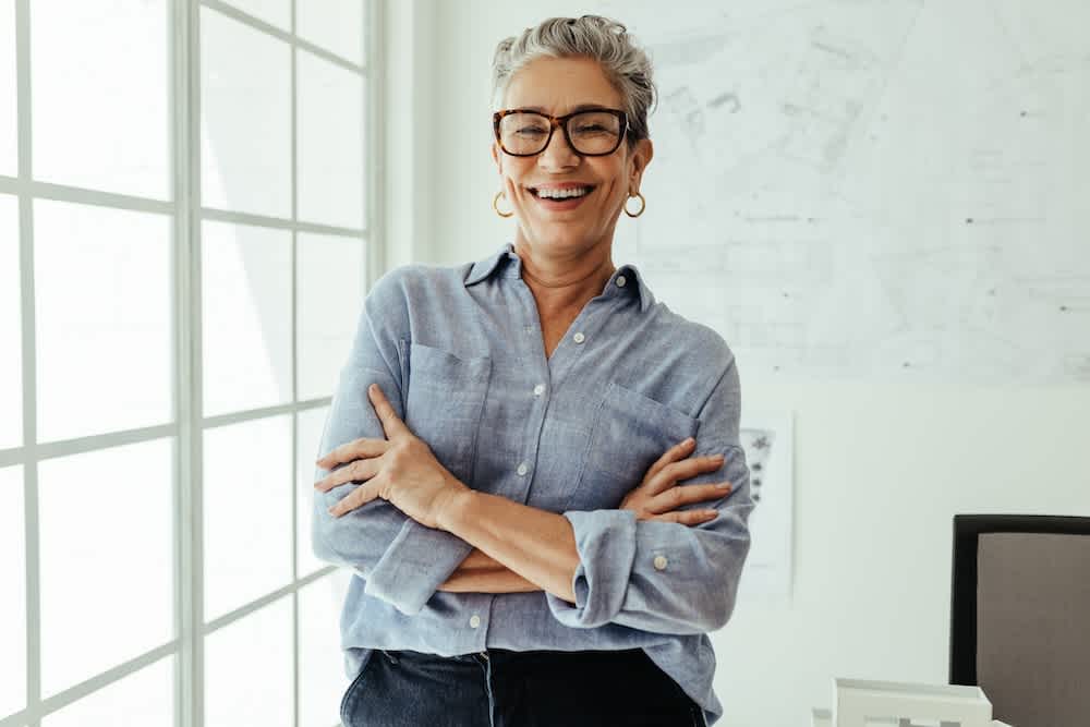 A smiling older woman with short gray hair, wearing glasses, hoop earrings, and a light blue shirt stands with arms crossed in a bright office with architectural plans on the wall behind her.