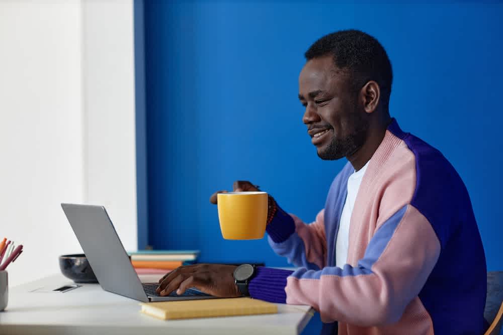 A man sits at a desk working on a laptop, smiling while holding a yellow mug. He wears a colorful sweater and watch, with notebooks and pencils on the desk, and a bright blue wall in the background.