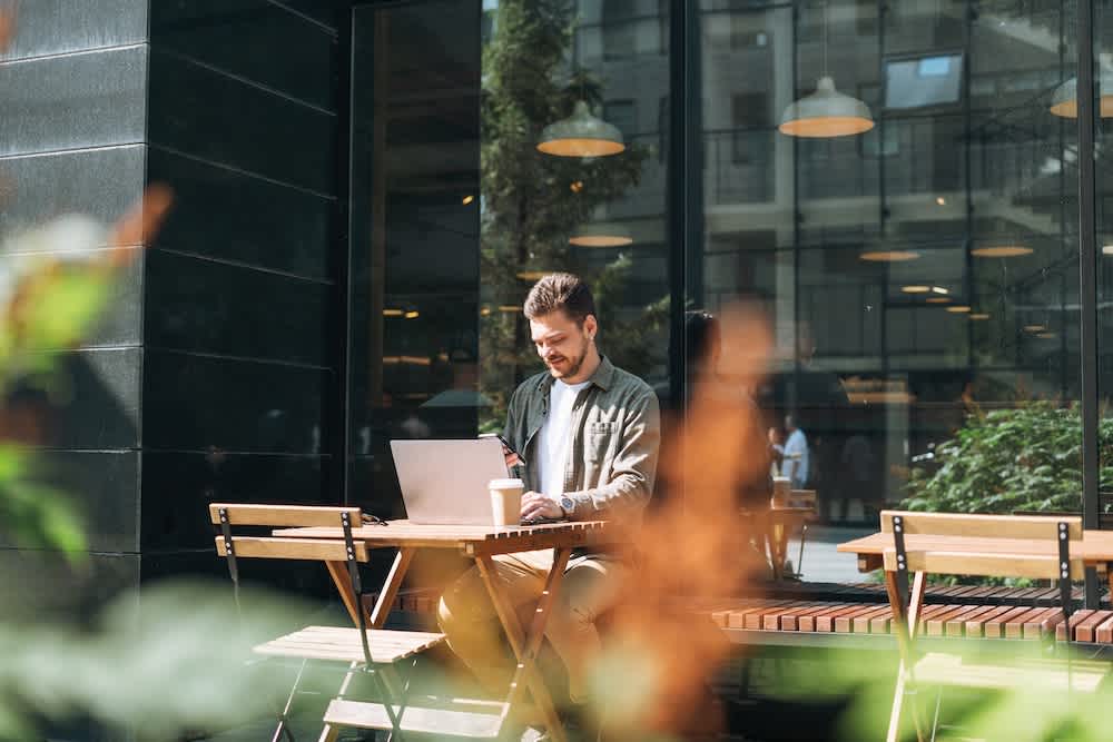 A man sits alone at an outdoor café table, working on a laptop with a coffee cup beside him. The background shows large windows, greenery, and a reflection of buildings and hanging lights.