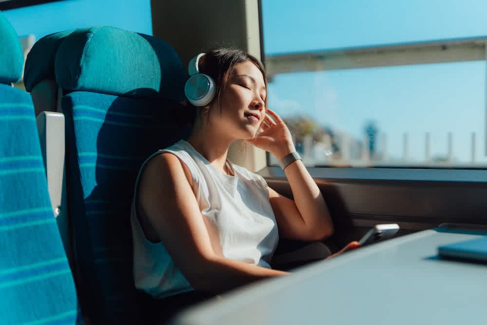 A woman wearing headphones sits by a window on a train, eyes closed and head tilted back, appearing relaxed while listening to music on her smartphone. Sunlight streams in through the window.