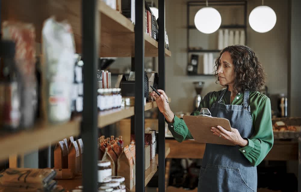 A woman wearing an apron and holding a clipboard checks inventory on shelves in a store or cafe, surrounded by various packaged goods and jars. The background features warm lighting and modern decor.