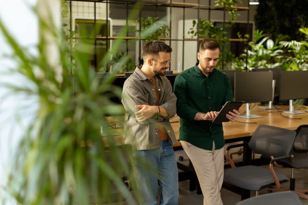 Two men stand in a modern office with plants and computers, looking at a tablet together. One man holds the tablet while the other watches attentively.