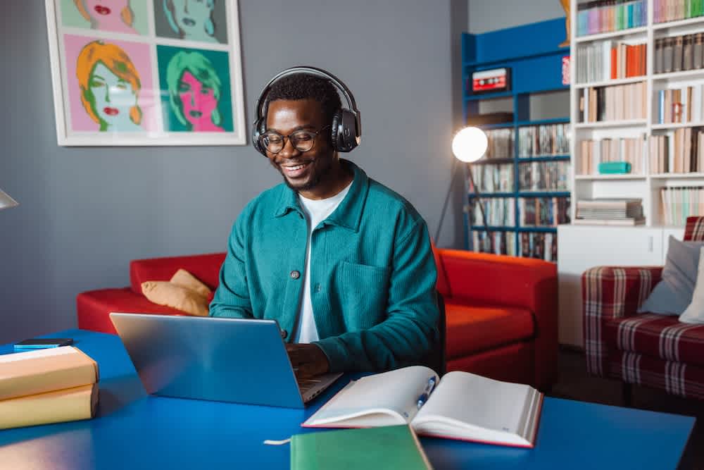 A person wearing headphones and a teal jacket smiles while using a laptop at a blue desk, with books and notebooks open nearby, in a colorful room with bookshelves, a red sofa, and pop art on the wall.
