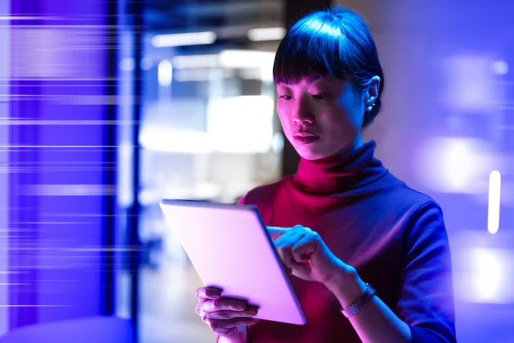 A woman in a red turtleneck uses a tablet in a modern, blue-lit office environment, with motion blur effects in the background suggesting technology and speed.