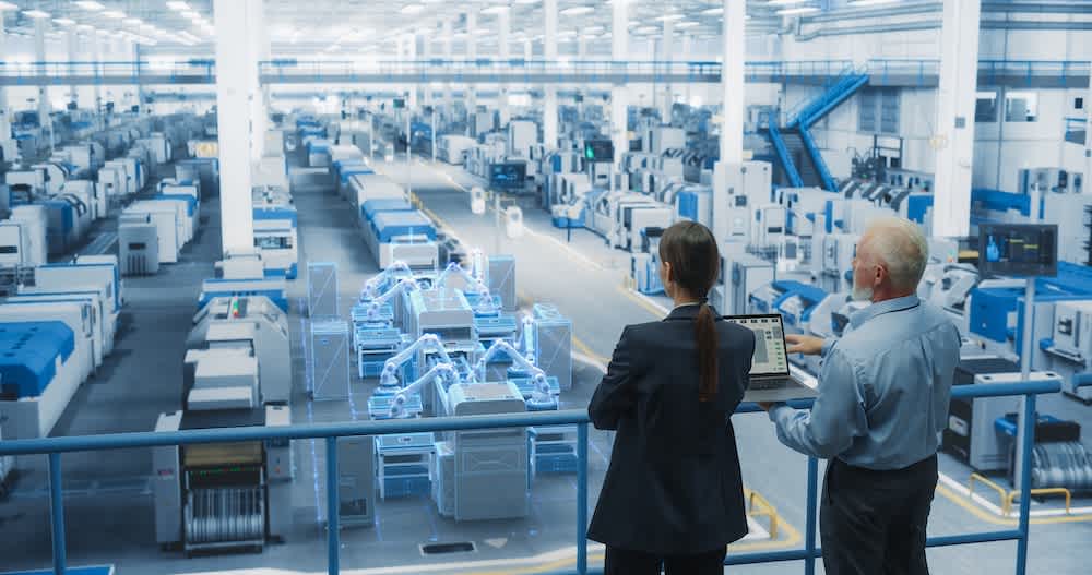 Two people overlook a large, modern factory floor with rows of machinery; a digital interface shows a holographic illustration of robotic arms, suggesting advanced automation and Industry 4.0 technology integration.