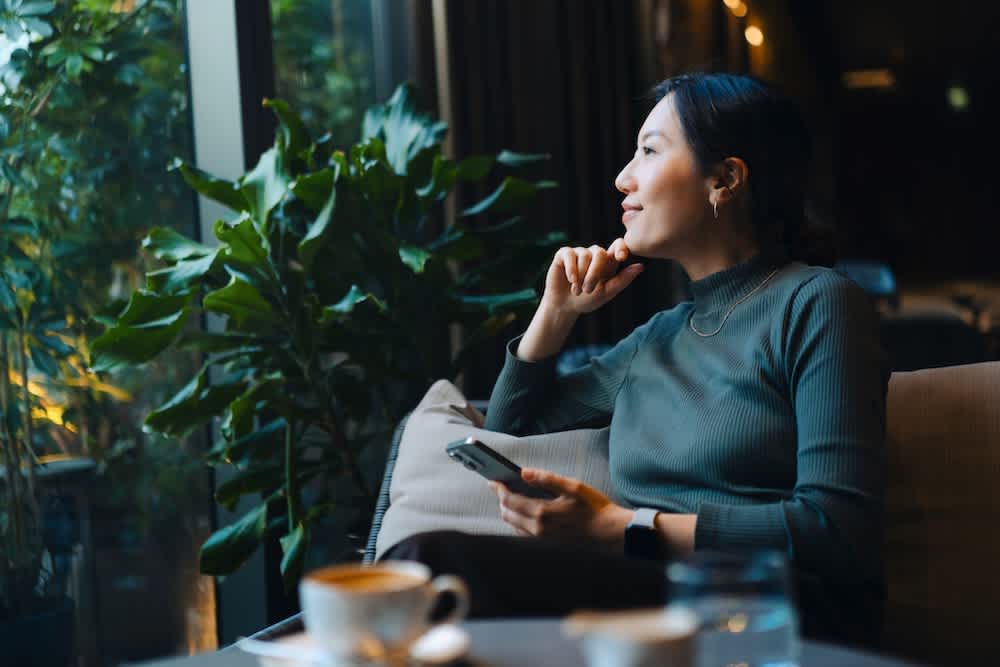 A woman sits on a couch by a large window, holding a smartphone and gazing outside thoughtfully. She is surrounded by green plants, with coffee and water on the table in front of her.