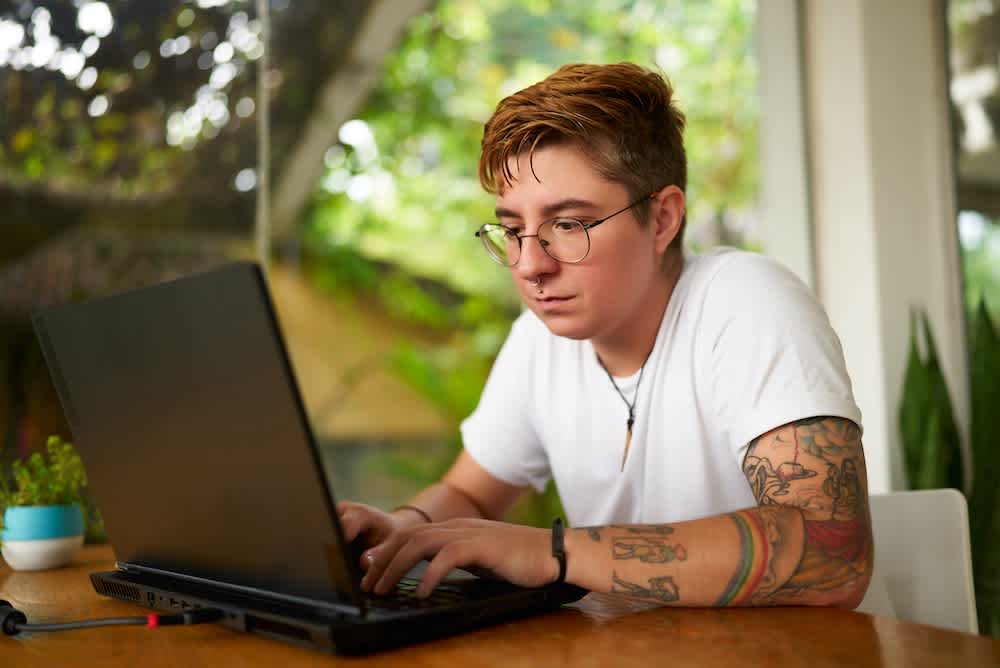 A person with short brown hair, glasses, and tattoos is typing on a laptop at a wooden table indoors, with green plants and a large window in the background.