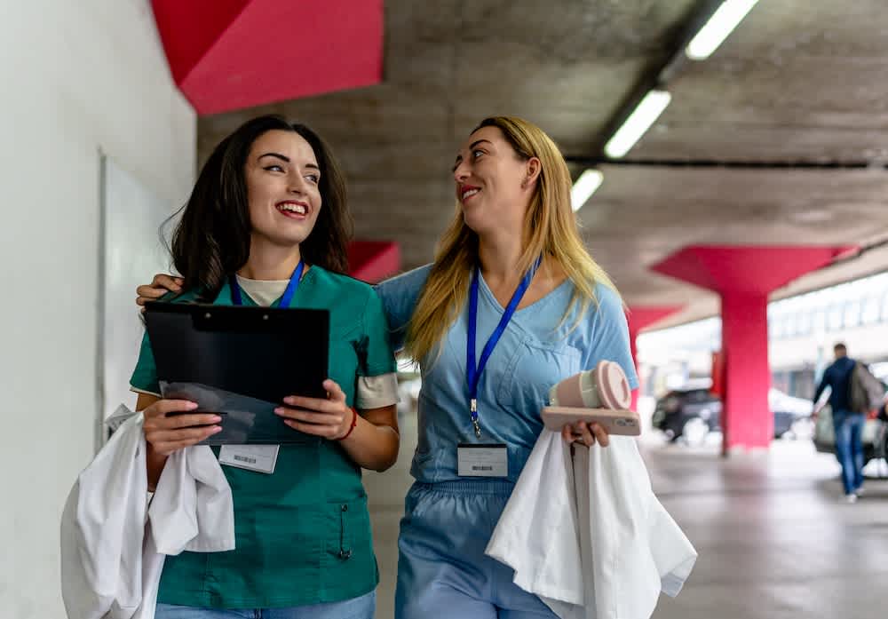 Two smiling healthcare workers in scrubs and name badges walk together under a covered walkway. One holds a clipboard, the other a coffee cup. Both appear cheerful and supportive, with one arm around the others shoulders.