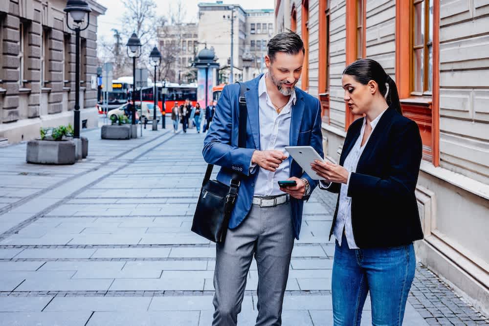 Two business professionals standing on a city sidewalk, discussing something on a tablet. The man is holding a phone and wearing a blue suit, while the woman holds the tablet and wears a black blazer and jeans.