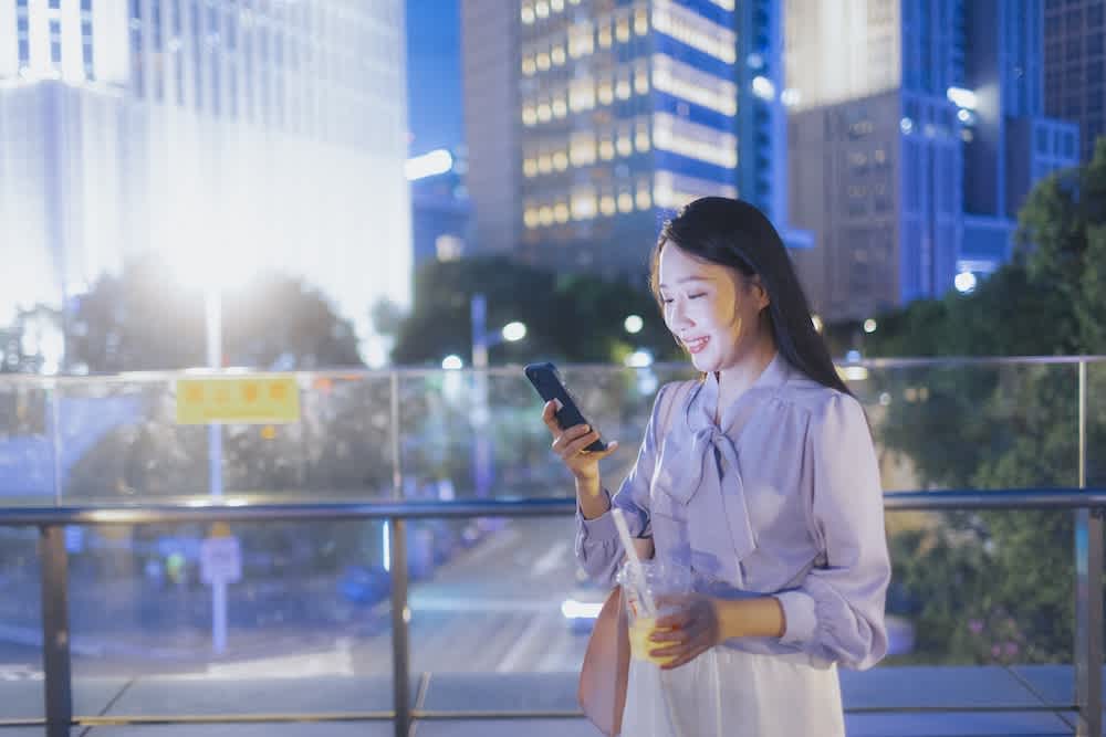 A woman standing outdoors at night in a city, smiling while looking at her phone and holding a drink, with tall buildings and bright lights in the background.
