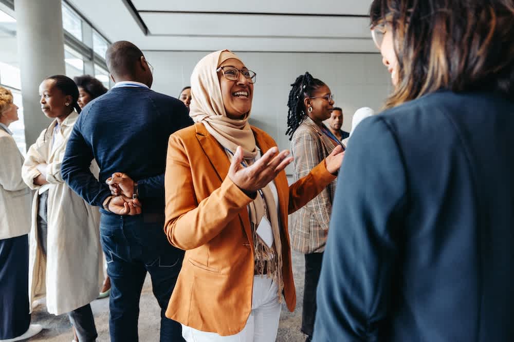 A group of people are standing and talking at a professional networking event. A woman in a tan blazer and headscarf is smiling and gesturing while speaking to another woman in the foreground.