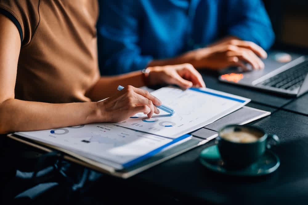 Two people review financial charts and graphs on paper at a table with a laptop and a cup of coffee, focusing on analyzing data with a pen.