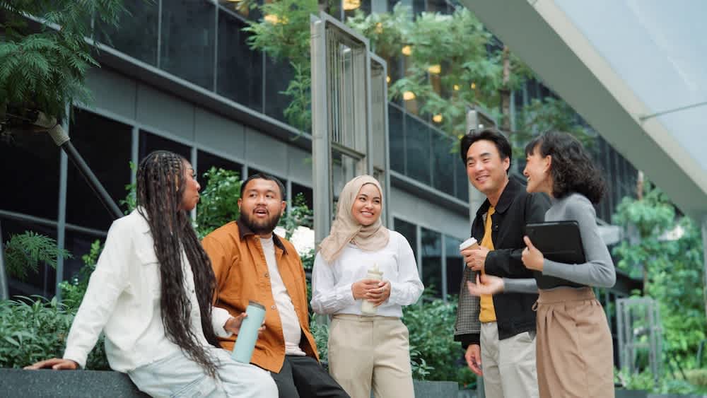 Five people, diverse in appearance and dress, stand and sit together outdoors by a modern building, smiling and talking. Some hold drinks and a tablet, surrounded by greenery and contemporary architecture.