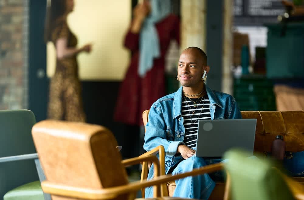 A person wearing a denim jacket and headphones sits on a chair with a laptop, smiling and looking to the side. Two people stand in the blurred background, talking. The setting appears to be a casual indoor space or café.