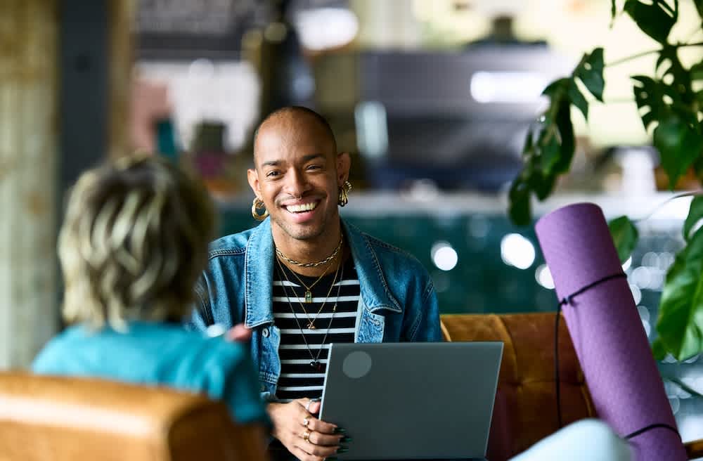 A person with short hair and multiple earrings smiles while holding a laptop and talking to another person in a casual, indoor setting with plants and a yoga mat nearby.