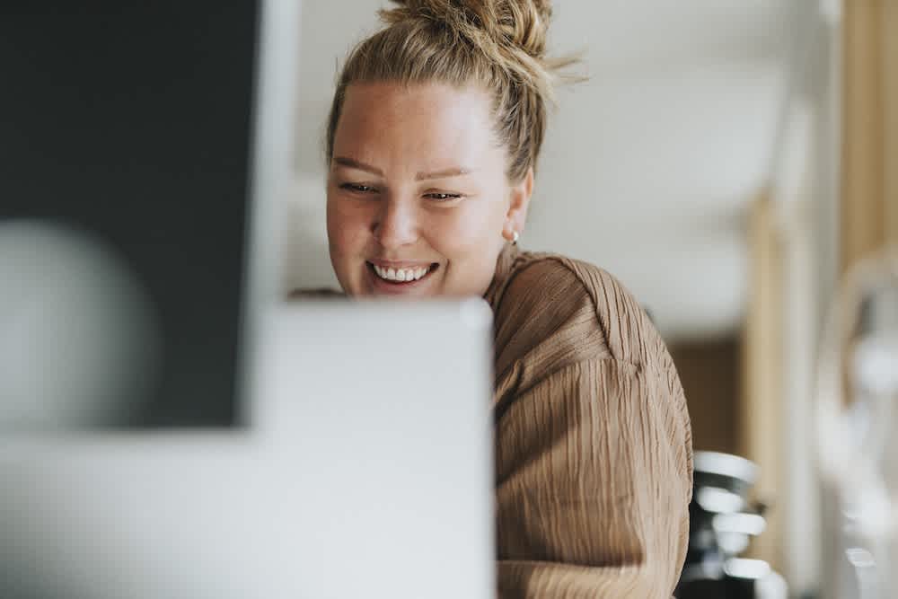 A smiling woman with her hair in a bun sits indoors, looking at a computer screen. She is wearing a brown top and appears to be enjoying what she sees on the monitor.