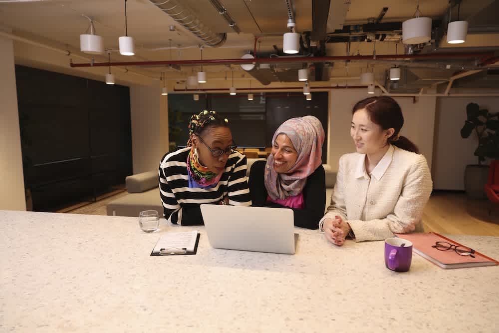 Three women sit together at a table in a modern office, smiling and looking at a laptop screen. One woman wears a striped shirt, another a hijab, and the third a white blazer with a pink folder and purple mug nearby.