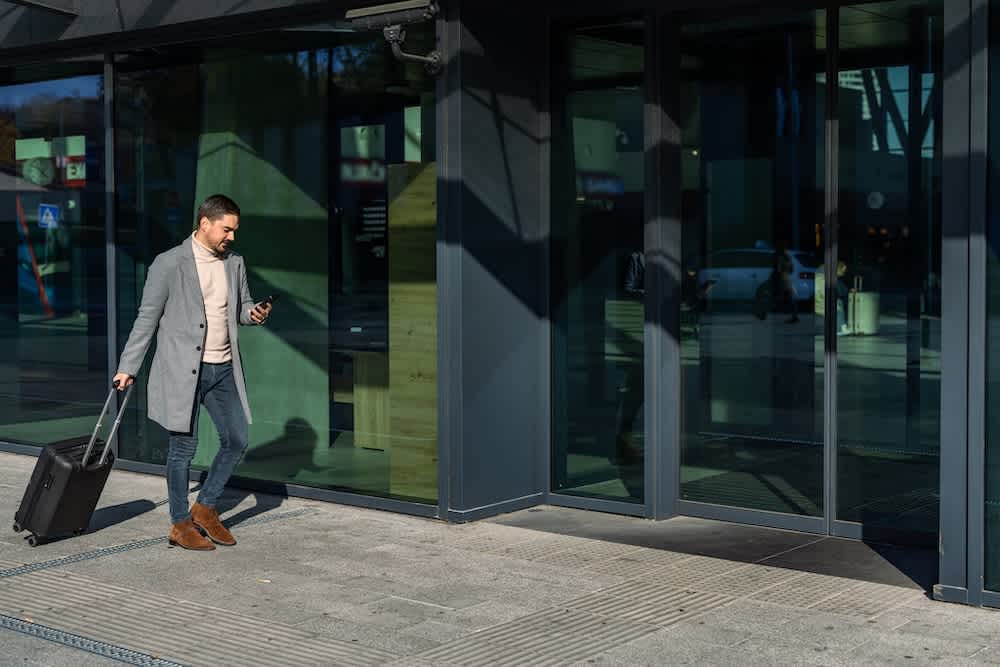 A man in a gray coat walks outside a modern glass building, pulling a black suitcase and looking at his phone.