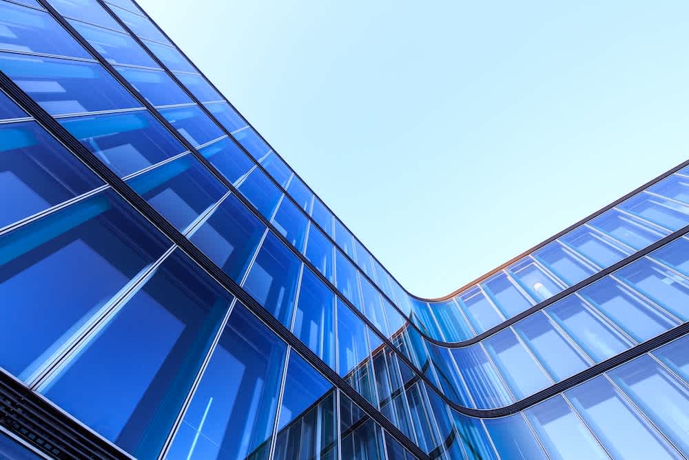 Looking up at the corner of a modern glass building with blue-tinted windows, reflecting the sky, under bright daylight. The architecture features smooth, curved lines and a sleek, contemporary design.