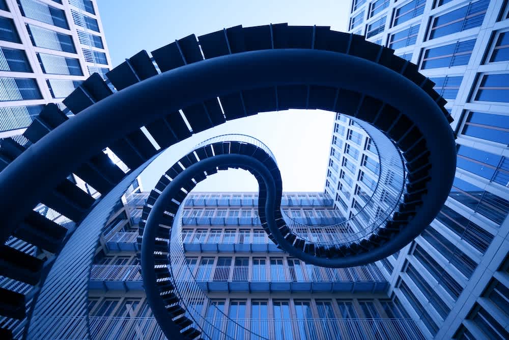 A spiral, looping outdoor staircase made of metal winds dramatically between modern, glass-sided office buildings, viewed from below against a bright blue sky.