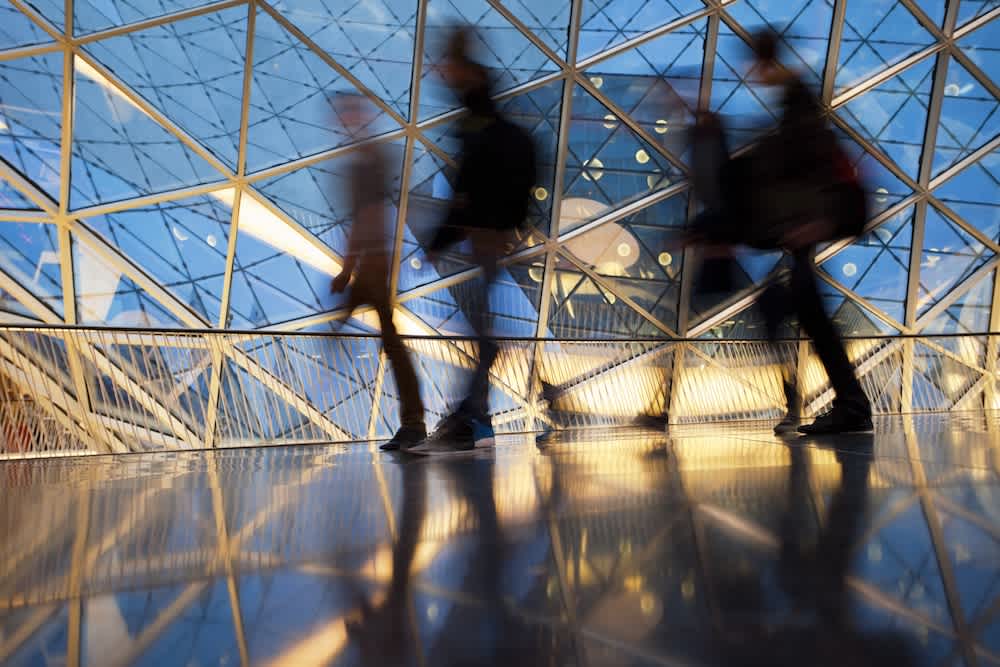 Blurred silhouettes of people walking inside a modern glass building with a geometric lattice pattern, reflections visible on the shiny floor, and blue sky in the background.