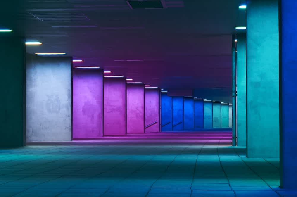 A modern tunnel with columns and walls illuminated by colorful lights in shades of pink, purple, blue, and teal, creating a gradient effect along the corridor’s length.