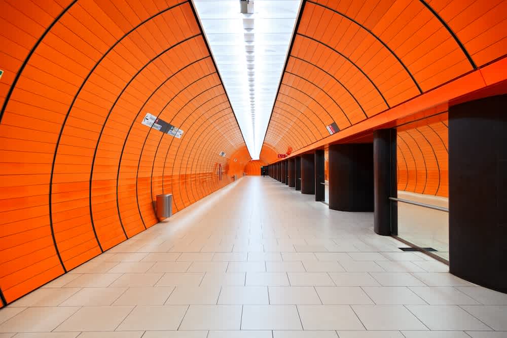 A modern, empty underground subway tunnel with bright orange curved walls, a white tiled floor, and black columns, creating a symmetrical, futuristic appearance. Signs and lights line the ceiling.