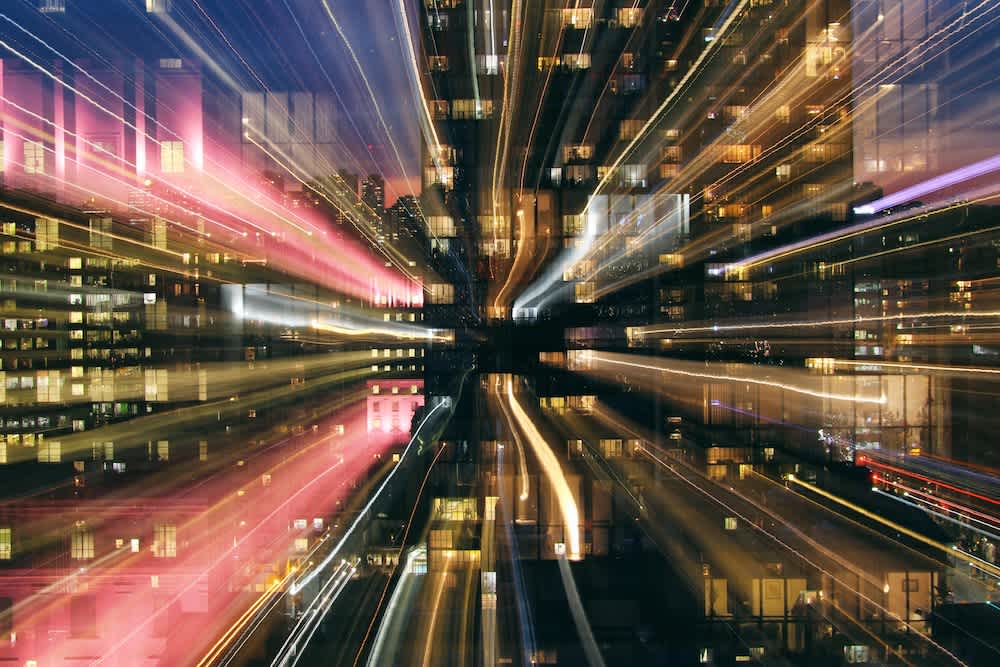 Nighttime cityscape with tall buildings and glowing windows. Light streaks radiate outward from the center, creating a dynamic, zooming effect with hues of pink, yellow, and blue.
