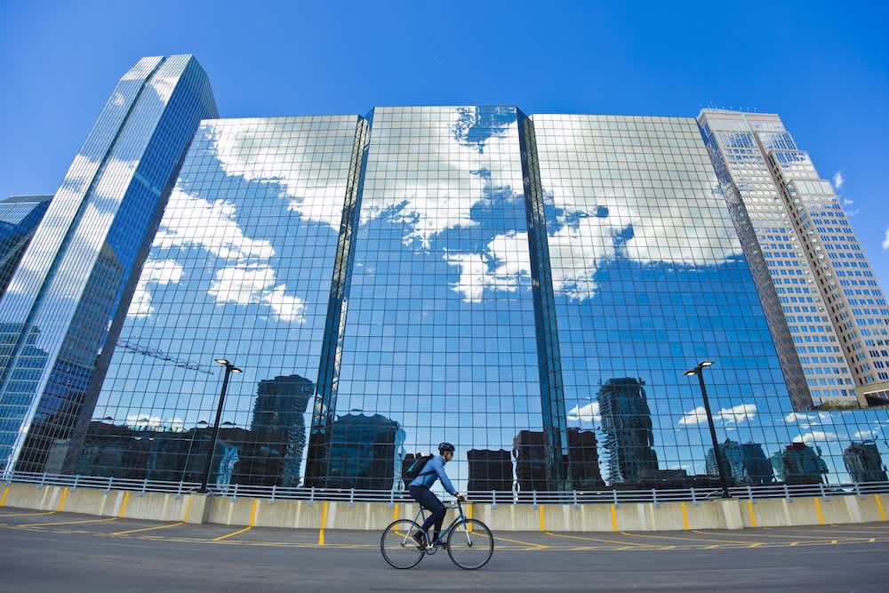 A person rides a bicycle in front of a tall glass building reflecting blue sky and clouds, with other city buildings also visible in the reflection.