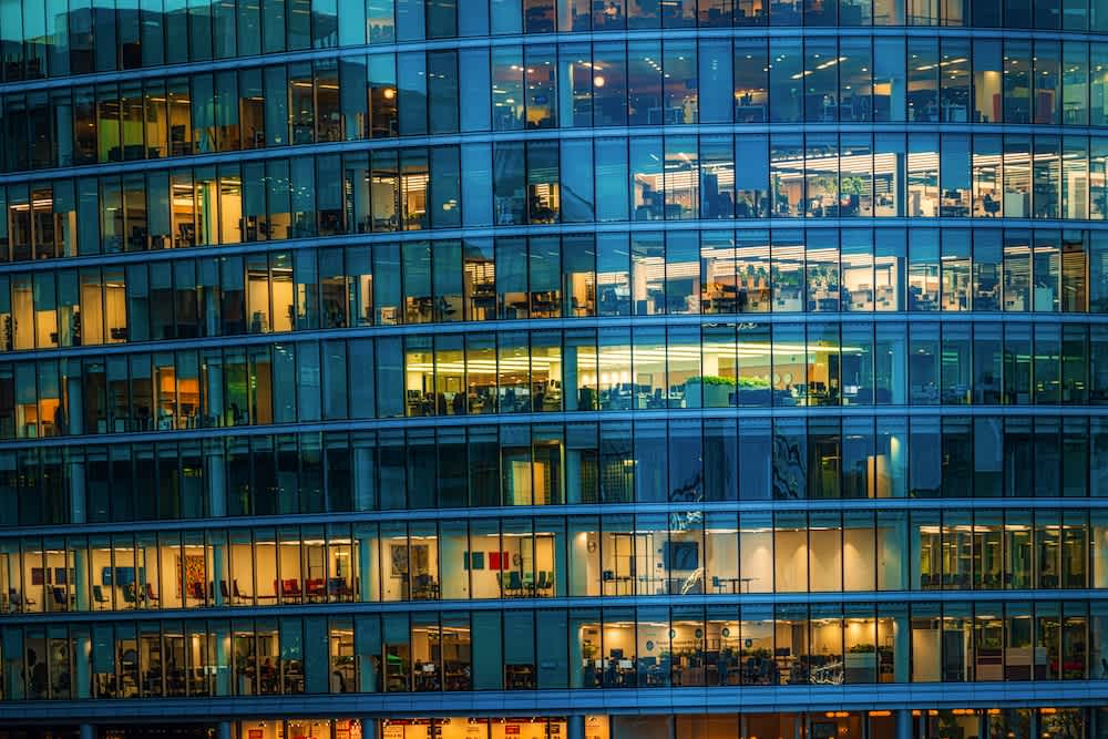 A close-up view of a modern glass office building at dusk, showing several floors with illuminated windows, revealing desks, chairs, and office equipment inside.