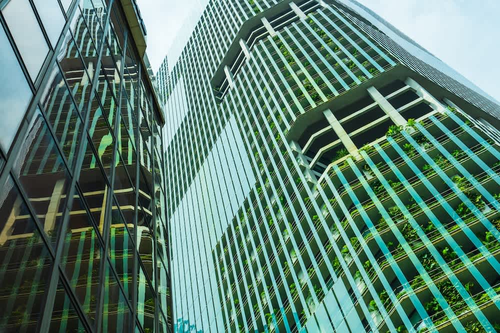 Tall modern buildings with glass facades reflecting each other, featuring green terraces and plants integrated into the architecture, set against a bright sky.