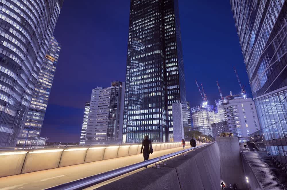 People walk on an elevated pathway surrounded by modern, tall office buildings at night, with the cityscape illuminated by artificial lights and cranes visible in the background.