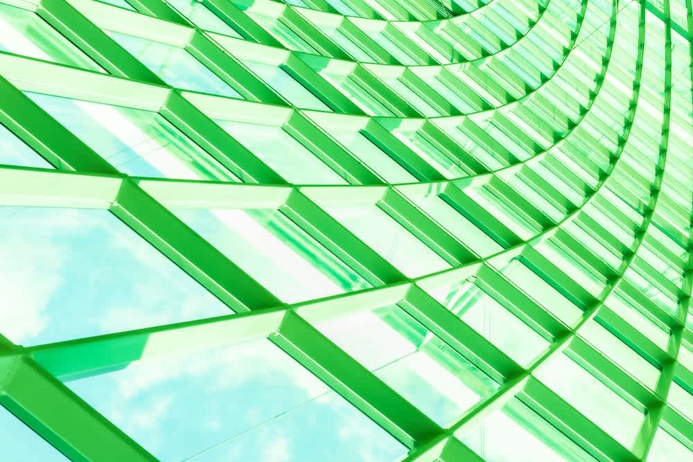 Abstract view of a modern glass building facade with green-tinted window frames, reflecting the sky and clouds. The windows form a wavy, geometric pattern that creates a sense of movement and depth.