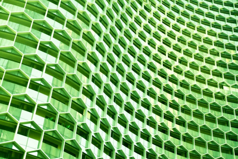 Close-up of a modern building facade featuring a repeating geometric pattern of hexagonal windows in varying shades of green, creating a textured, honeycomb-like appearance.