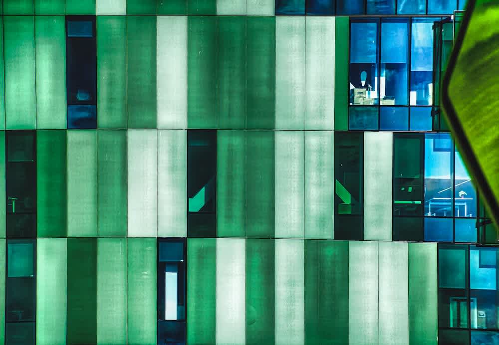 A modern building facade with alternating vertical green and translucent panels, and blue-tinted windows revealing office interiors. A green geometric shape is visible in the foreground on the right side.