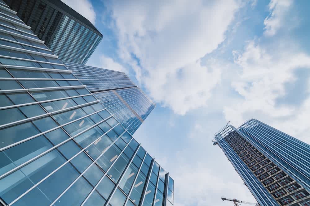 Skyscrapers with glass facades rise toward a blue sky with scattered clouds. A building under construction with cranes is visible on the right side of the image. The perspective is from ground level looking up.
