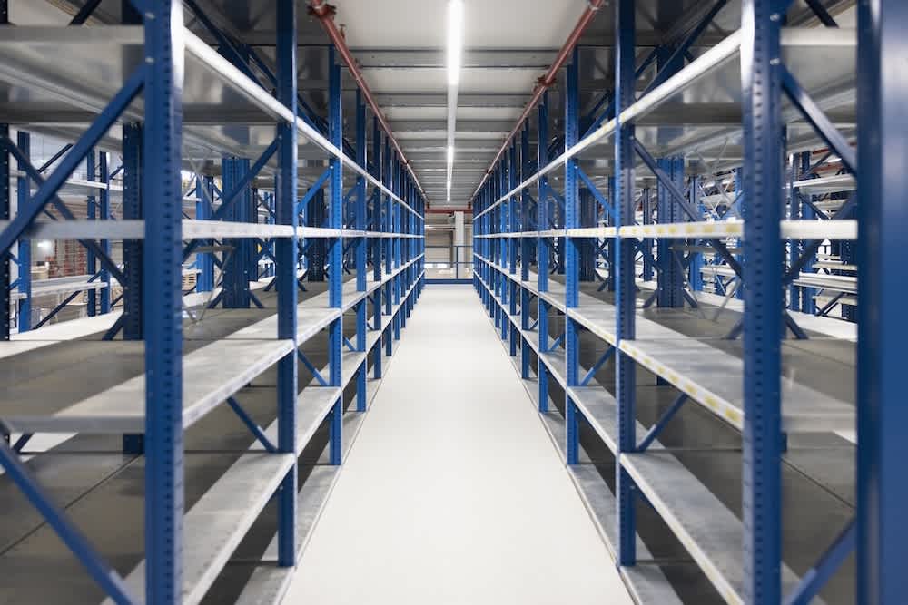 A straight-on view of multiple rows of large, empty metal shelving units in a brightly lit warehouse or storage facility, creating a symmetrical corridor between the racks.