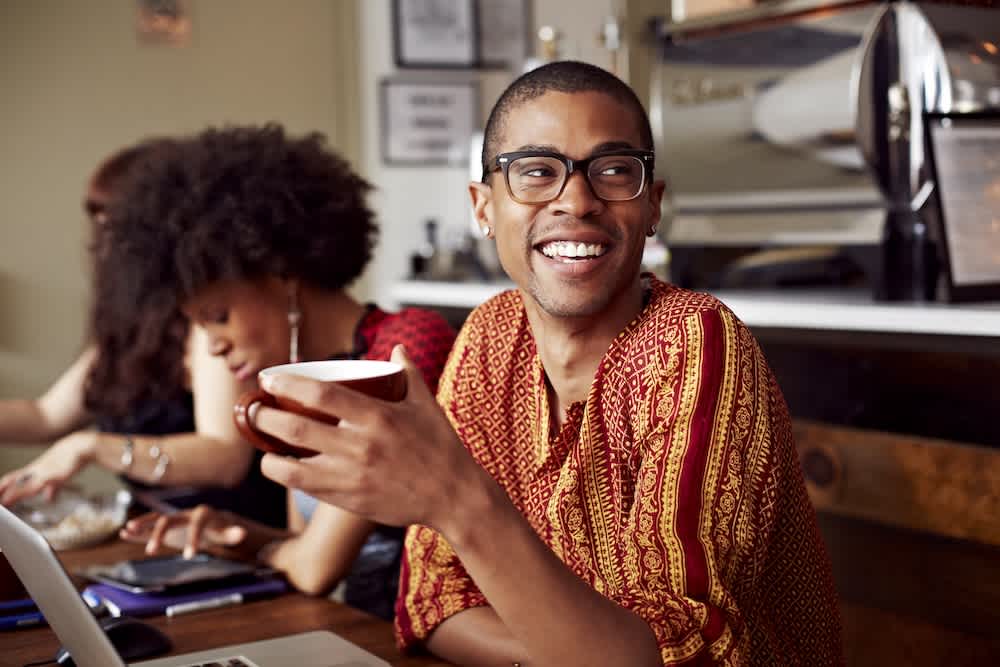 A smiling person wearing glasses and a patterned shirt holds a coffee cup while sitting at a café table; another person with curly hair works beside them on a laptop.