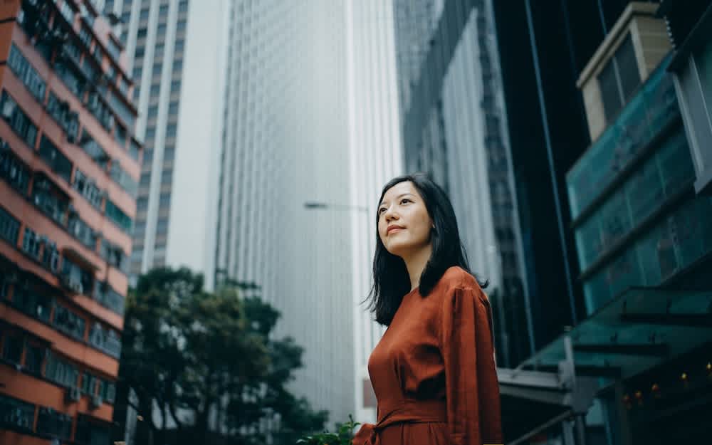 A woman in a rust-colored dress stands outdoors, looking up and smiling, surrounded by tall modern skyscrapers and a red apartment building in an urban cityscape.
