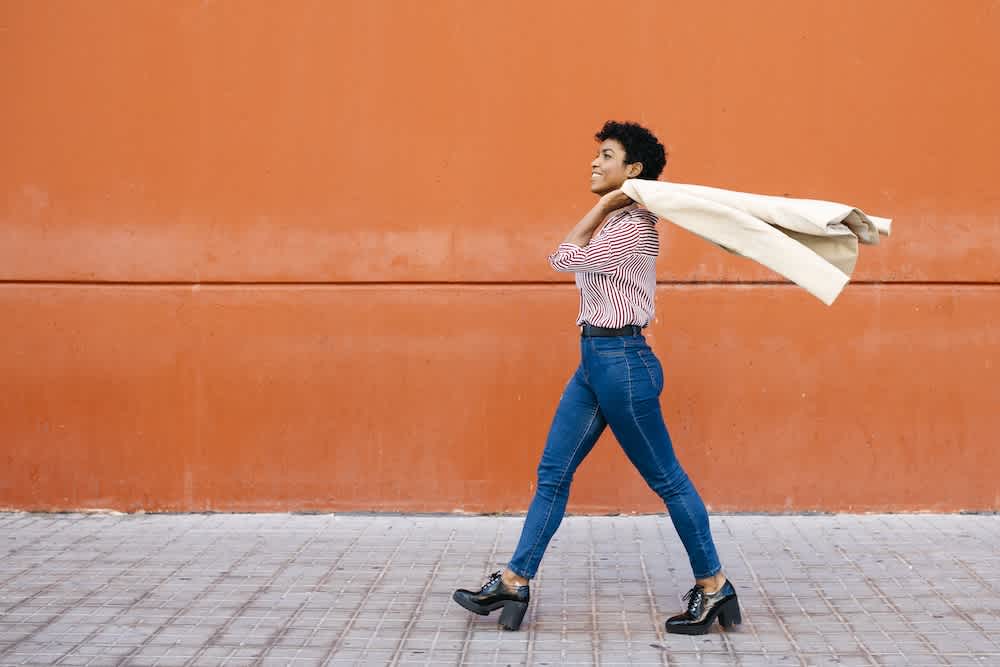 A woman in jeans and heeled boots walks confidently along a tiled sidewalk, smiling and holding a beige coat over her shoulders, with an orange wall in the background.
